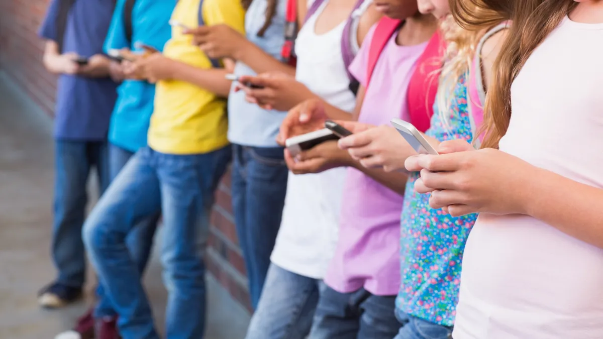 A row of students standing and leaning against a brick wall. In their hands are cellphones